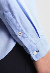 Camicia button-up azzurro chiaro con maniche, caratterizzata da una finitura testurizzata, con bottoni bianchi e accenti contrastanti in rosso e blu.