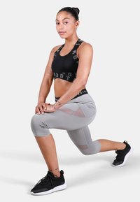 Juoda sportinė liemenėlė su spausdintu elastingu diržu, pilkos spalvos capri tipo legginai su tinklinėmis detalėmis, juodi sportiniai batai. Modelis atsidūręs pusiau pritūpimo pozicijoje.