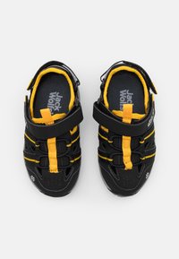 Mustat säädettävät kengät keltaisilla yksityiskohdilla, joissa on teksturoitu pinta, hengittävä verkko ja varma remmikiinnitys helppoa käyttöä varten.
