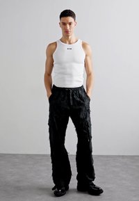 Un débardeur blanc côtelé et un pantalon large noir déchiré, présentant une coupe ample et des accents texturés, associés à des chaussures noires imposantes.