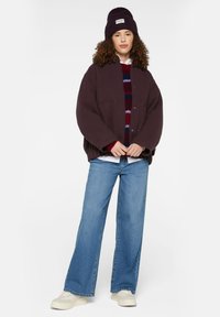 Übergroße dunkelviolette Jacke mit zwei frontalen Taschen, lässig über einem gestreiften Pullover getragen. Hellblaue weite Jeans und beigefarbene klobige Sneakers.