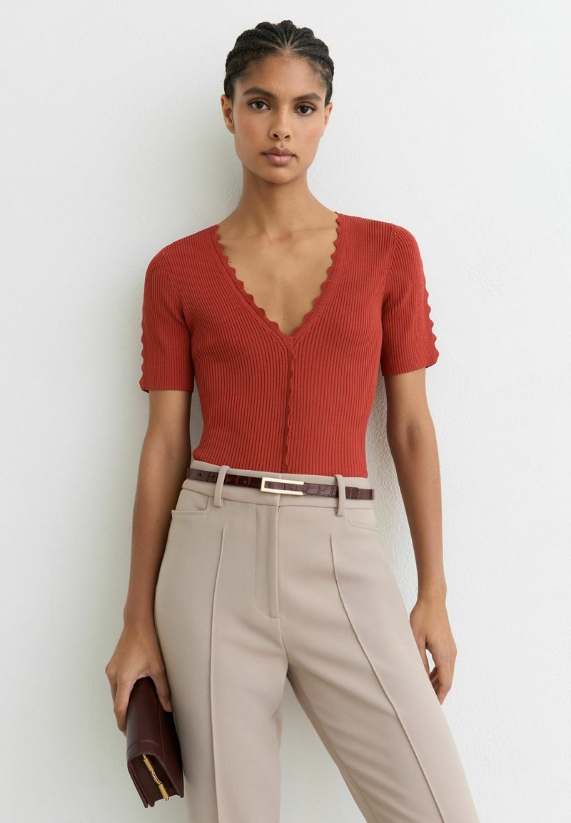 Vrouw draagt een roestrode, geribbelde top met geschulpte randen, beige broek met hoge taille, bruine riem, houdt een bruine clutch vast tegen een witte muur.