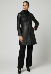 Kvinna i svart figurnära läderrock med knappar, svart polotröja, vida byxor och svarta stövlar med klack, stående mot en enfärgad bakgrund.