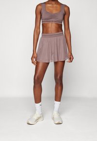 Bralette sportivo marrone con fronte arricciato, abbinato a una mini gonna a campana dello stesso colore. Indossato con calze bianche e scarpe da ginnastica beige.