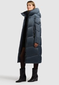 Dolga temno modra puffer jakna z visokim ovratnikom, zadrgo in diagonalnim kvadratnim vzorcem. Nosi se preko črne majice in v paru s črnimi klino čevlji.