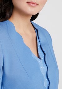 Light blue scalloped jacket with a smooth texture, layered over a light blue polka-dotted top. Features a deep V-neck design.