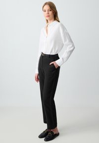 Weißes Leinenhemd mit langen Ärmeln, schwarze maßgeschneiderte Hosen mit Taschen und schwarze Slipper mit goldenen Hardware-Details. Einfacher, strukturierter Look.
