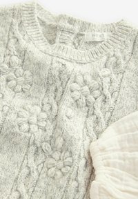 Pull en tricot gris avec des motifs floraux en relief et un col côtelé ; à côté se trouve un morceau de tissu texturé de couleur claire.