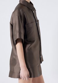 Koszula z krótkim rękawem, z kołnierzykiem, z ciemno-brązowego satynowego materiału. Posiada zapięcie na guziki z przodu oraz kieszonkę na piersi. Gładka tekstura z delikatnym połyskiem.