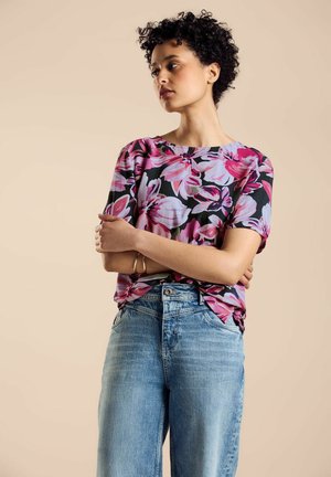 Schwarze Bluse mit kurzen Ärmeln, die ein lebhaftes rosa Blumenmuster aufweist, kombiniert mit hellblauen, hochtaillierten Jeans. Strukturiertes Material und lockere Passform.