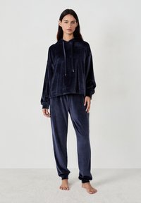 Kobieta stoi boso, ubrana w granatowy aksamitny hoodie i pasujące do niego aksamitne spodnie dresowe na jednolitym bia łym tle.