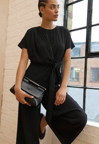 Schwarzer strukturierter Jumpsuit mit kurzen Ärmeln und einer Frontschnürung. Getragen mit einer schwarzen Clutch. Geflochtener Haarschnitt, steht nahe einem Fenster.