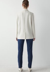 Blazer blanc texturé avec une seule fente au dos, associé à un jean bleu foncé ajusté. Le modèle porte des talons crème. Design minimaliste, lignes épurées.