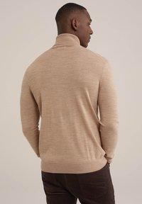 Beige kötött pulóver magas gallérral, testhez simuló fazonnal és bordázott aljjal. Sötét barna nadrággal viselve, amely textúrát és puhaságot mutat.