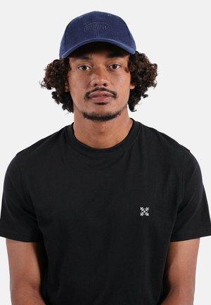 Homme aux cheveux bouclés portant une casquette bleue et un t-shirt noir avec un petit logo blanc sur la poitrine, faisant face à l'avant sur un fond uni.