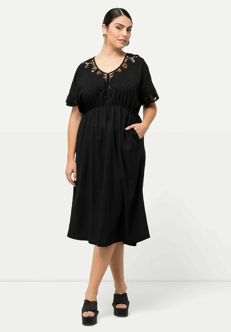 Vestido maxi negro con detalles de encaje en el escote y mangas cortas, con una cintura ceñida y bolsillos laterales. Se lleva con zapatos de plataforma negros.