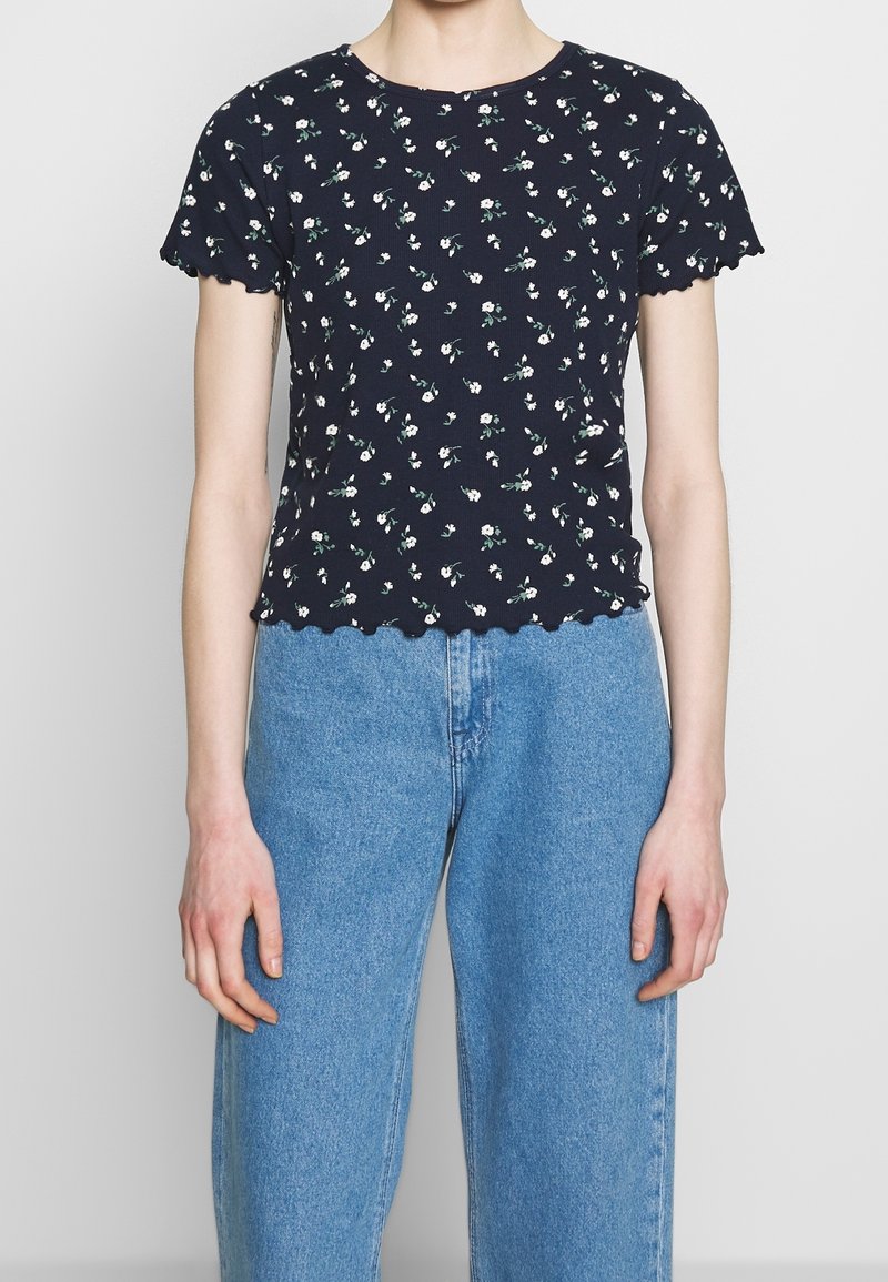 T-shirt navy con stampa floreale, a maniche corte e bordi ondulati, caratterizzato da piccoli fiori bianchi e foglie verdi, realizzato in morbido cotone.