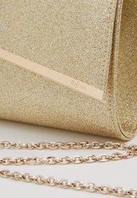 Pochette dorée pailletée avec bandoulière chaîne et bande beige avec le nom de la marque "Mascara" sur le côté.