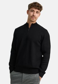 Musta fleece-pullover, jossa on puolivetoketjukaulus, ribbikuteet hihansuissa ja helmassa. Hihassa on sileä pinta ja rento istuvuus. Yhdistetty harmaisiin housuihin.