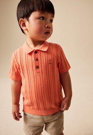En ung dreng iført en koralfarvet kortærmet strikket polo og beige shorts, der kigger mod højre mod en ensartet lys baggrund.