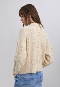 Pull crème en maille torsadée avec col montant, orné de perles transparentes. Coupe ample avec poignets et ourlet côtelés, porté avec un jean bleu.