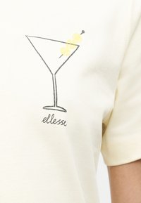 Περίγραμμα ποτηριού μαρτίνι με δύο κίτρινες ελιές σε καλαμάκι, με το κείμενο «ellesse» από κάτω, σε φόντο από μπεζ ύφασμα.