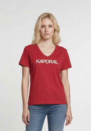 Donna bionda che indossa una T-shirt rossa con scollo a V e la scritta "KAPORAL" e jeans blu, in piedi davanti a uno sfondo chiaro e uniforme.