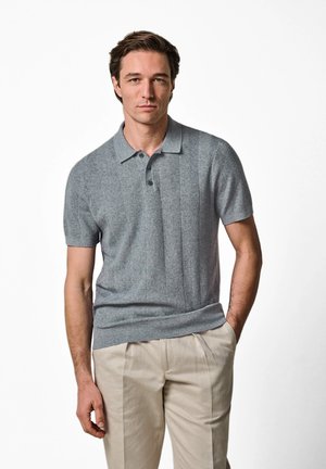 Mand iført en kortærmet grå strikket poloshirt og beige bukser, stående med den ene hånd i lommen mod en hvid baggrund.