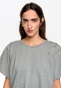 Szary t-shirt z krótkim rękawem, z bufkami na ramionach i metalowymi ćwiekami wzdłuż szwu. Wykonany z miękkiej bawełnianej mieszanki, z okrągłym dekoltem.