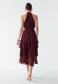 Burgundi spalvos tiered maksi suknelė, pagaminta iš lengvos medžiagos, su halter tipo kaklu ir juosmeniu apjuostu diržu. Sklaidomi sluoksniai sukuria plazdantį siluetą.