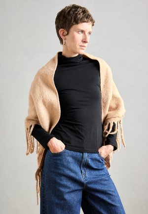 Sciarpa di lana beige con bordo frangiato appoggiata su un dolcevita nero. La modella si trova con le mani in tasca, su uno sfondo neutro.
