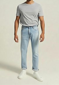 Homme portant un t-shirt gris clair, un jean bleu clair retroussé et des baskets blanches, debout devant un fond uni clair.