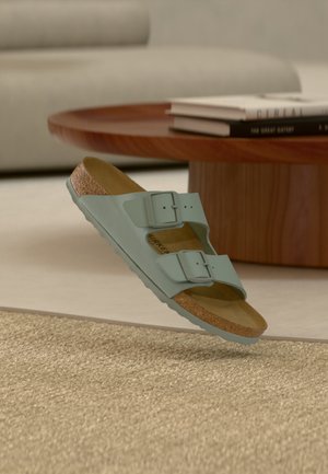 Sandale bleu clair à deux brides avec semelle en liège flottant au-dessus d'un tapis beige texturé, table basse en bois et livres en arrière-plan dans le salon.