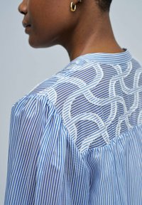 Blau-weiß gestreifte Bluse mit einem transparenten Rücken, der mit gestickten kreisförmigen Mustern verziert ist, sowie gerafften Details an den Schultern.