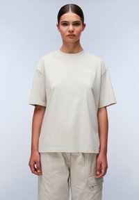 Beige bomulds t-shirt med en løs pasform, rund hals og korte ærmer; har et diskret logo på brystet, sammen med beige cargo-bukser.