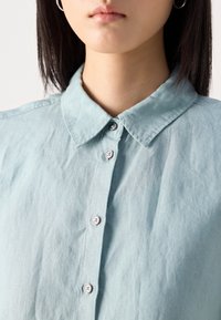 Gros plan d'une personne portant une chemise bleu clair boutonnée avec un col et des boucles d'oreilles argentées en forme d'anneau, montrant le bas du visage et le cou.