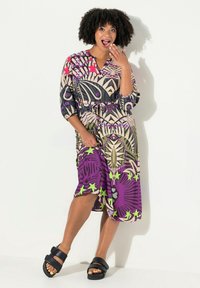 Bunt gemustertes Kleid mit geometrischen Mustern, V-Ausschnitt, tailliert und dreiviertel Ärmeln. Kombiniert mit schwarzen Sandalen.