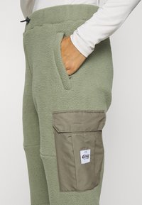 Fleece pants in soft green with textured fabric, featuring side pockets and a patch pocket in a darker green, branded label visible.