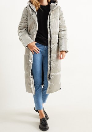 Femme portant un long manteau matelassé gris clair avec capuche, top noir, jean bleu et mocassins noirs, debout devant un fond uni.