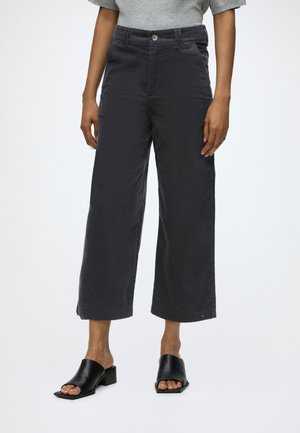 Personne portant un pantalon large noir raccourci, un haut gris et des mules à talons ouvertes noires sur fond blanc.