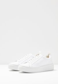 Ett par vita låga sneakers med tjocka sulor och snören på en vit bakgrund, vinklade något åt höger.