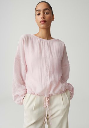 Femme portant un chemisier rose clair à manches longues avec un ourlet à cordon de serrage et un pantalon blanc, debout les mains dans les poches devant un fond uni.