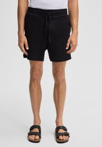 Zwarte textuur short met een elastische tailleband en trekkoord, zijzakken en een relaxte pasvorm. G gedragen met zwarte sandalen.