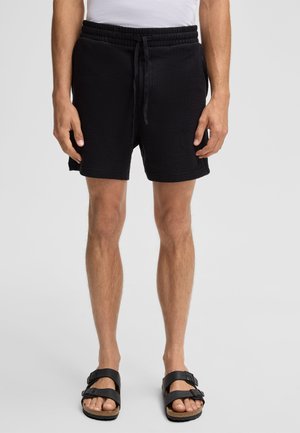 Schwarze strukturierte Shorts mit einem elastischen Bund und Kordelzug, seitlichen Taschen und einem lässigen Schnitt. Getragen mit schwarzen Sandalen.