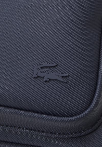 Un matériau noir texturé met en avant un logo de crocodile en relief bien visible. Le design comprend des bords cousus et une finition lisse.