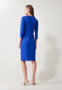 Robe ajustée bleue avec des manches trois-quarts, ornée d'une bordure en dentelle à l'ourlet. Tissu lisse et silhouette cintrée, longueur cheville.