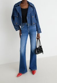 Giacca in denim cropped con grande colletto, bottoni doppi; abbinata a jeans blu a zampa d'elefante, tacchi a punta rossi e una piccola borsa nera.