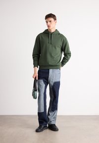 Felpa color verde oliva con cappuccio e lacci, abbinata a pantaloni in denim a pezzi in tonalità chiare e scure, e scarpe da ginnastica nere. Tiene una borsa piccola.