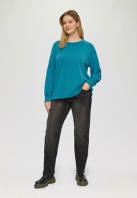 Türkisfarbener Sweatshirt mit langen Ärmeln und rundem Ausschnitt, kombiniert mit schwarzen Jeans und klobigen schwarzen Stiefeln, vor einem schlichten Hintergrund stehend.