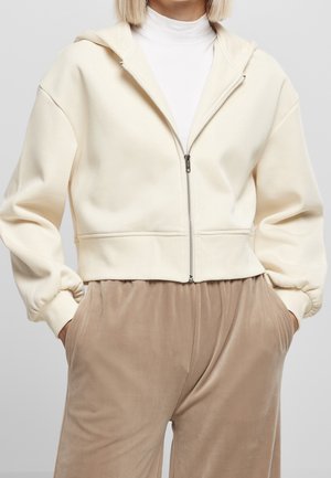 Personne portant un sweat à capuche crème zippé sur un col roulé blanc et un pantalon ample beige, les mains dans les poches, sur un fond uni.